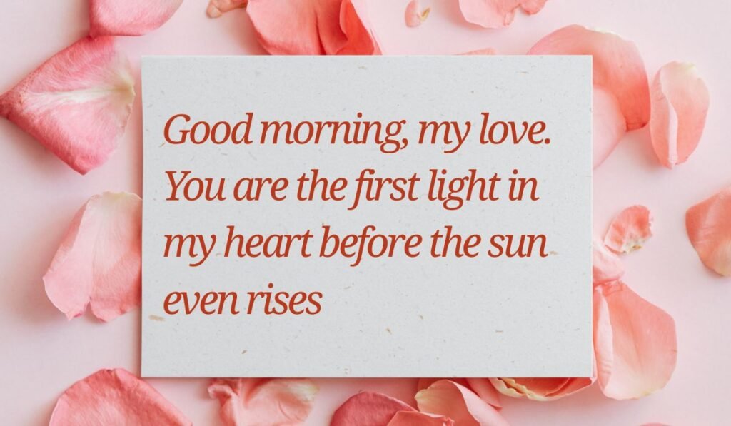Elegant white card surrounded by soft pink rose petals on a light pink surface, displaying romantic good morning messages for her: "Good morning, my love. You are the first light in my heart before the sun even rises".